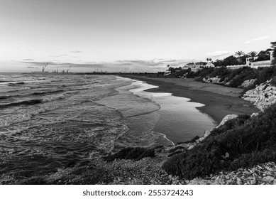 Black and white coastal landscape with gentle waves, a sandy beach, rocky shoreline, houses, and distant industrial cranes. A blend of natural beauty and urban elements for artistic or editorial use. - Powered by Shutterstock