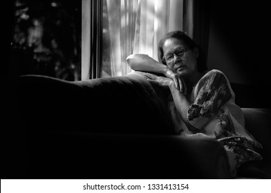 Black And White Dramatic Low Key Portrait Of 70s  Asian Elderly Woman Looking To The Camera,eyes Contact,rim Light Upon Her Body.The Light And Shadow Of Morning Climate Background.Sad Elderly Concept.