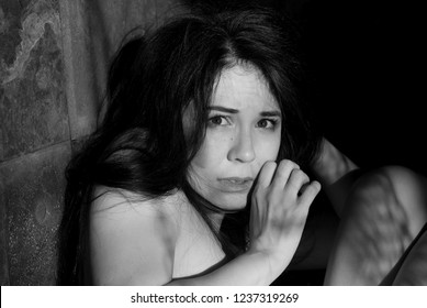 Black and white emotional portrait of a frightened and tense young woman looking out the window and covering her face with her hands back against a rusty wall in a dark room. Negative emotions.