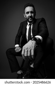 Black And White. Friendly Smiling Man In Modern Office Suit Sits Holding Hand On His Knee, Looking Down At Camera, Ready To Help Over Dark Background