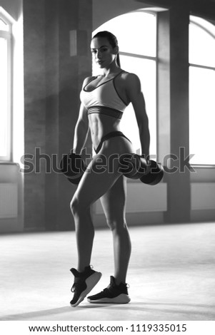 Black and white frontview of athletic sporty woman posing with dumbbells in spacy gym with panoramic windows. Having strong, fit body with heatlthy tanned skin and muscles. Doing fitness exercises.