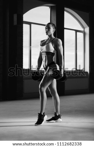 Black and white frontview of athletic woman posing with dumbbells in spacy gym with panoramic windows. Having strong, fit body with heatlthy tanned skin and muscles. Doing fitness exercises.