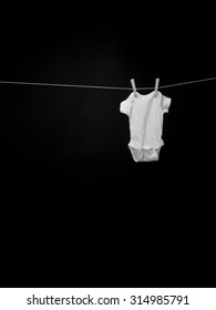 Black And White Image Of A Baby Onesie Hanging On A Clothesline In Front Of A Black Background