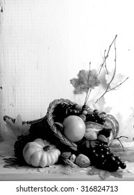 Black and white image of fall cornucopia containing fruits and vegetables and nuts with autumn leaves scattered around with rustic wood background. Vertical composition with copy space in upper left.