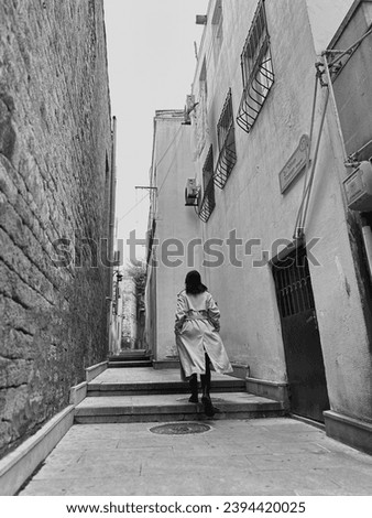 Black and white image: girl in white long coat is walking through the old ancient town alone. Woman in fashion coat in motion among white walls back view. Street walker. Beautiful girl alone is going