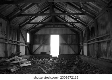 A black and white image of the inside of a large and abandoned metal walled barn.  A door in the far wall is brightly by the sunshine outside.