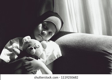 Black And White Image Of Patient Kid Lie Down On Couch With Doll.Girl Cover Her Head With Headscarf.Kid Look Sad,tired And Sick.Concept Of   Childhood Cancer Awareness.Selective Focus.