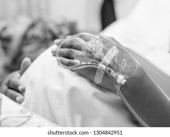A black and white image of a pregnant lady lying in a hospital bed holding her tummy, with a close up on the drip in her hand
