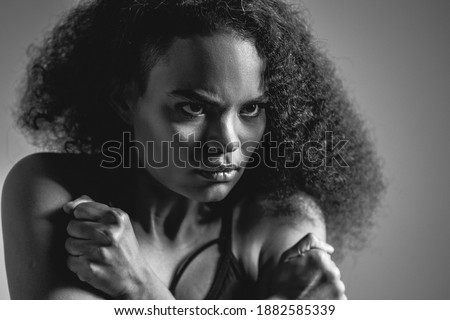 Black and white image. Self-defense, fearful African American girl standing covered with hands in black bare shoulder top isolated on grey background. Human emotions, facial expression concept.