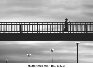 Black and white image of silhouette woman in protective mask and using mobile phone while walking on the overpass after get off work against sunset sky background in the city - Powered by Shutterstock