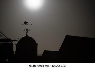 Black and white image of weather vane on top of cuppola roof with sun in the background