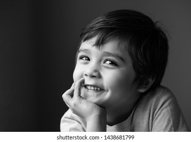 Black And White Indoor Portrait Of Adorable Little Boy Sitting Alone And Looking At Camera With Smiling Face, Low Key Head Shot Happy Kid Relaxing At Home, Positive Child With A Happy Face