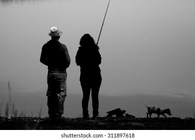 Black And White Landscape With Couple And Small Dog Fishing Near Small Lake 