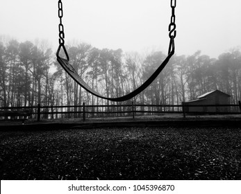 Black and white of a lone empty swing on an dreary cold foggy morning with a barn and a wooden fence in the background.