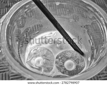 ​Top-down black and white macro shot of refreshing iced coffee with hollow tube ice cubes and a black straw inside a clear plastic cup, featuring detailed condensation droplets.