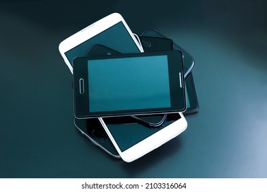 Black And White Mobile Smartphones On Dark Background. Mobile Phones In Stack On Dark Table, Top View. Set Of Contemporary Smartphones. Wireless Communication Technology And Mobility Concept