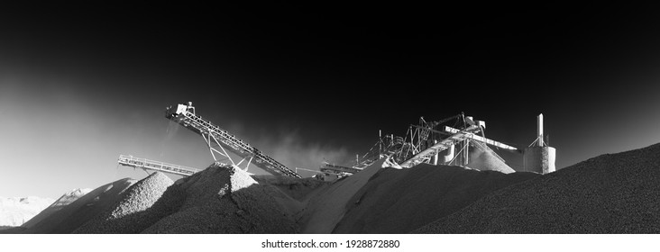 Black and white panorama of rock crushing equipment at a mining plant, working in a cloud of stone dust on the background of impenetrable black sky.  Mining industry.