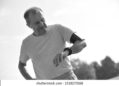 Black And White Photo Of Active Senior Man Standing With Hands On Hips While Checking Watch In Park