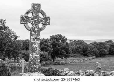 A black and white photo of an ancient Celtic cross in a peaceful rural cemetery, surrounded by stone walls, trees, and distant rolling hills under a cloudy sky. - Powered by Shutterstock