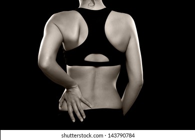 A black and white photo of an athletic woman holding her lower back as if experiencing pain. Photographed in the studio. Isolated on a black background. - Powered by Shutterstock