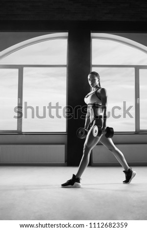 Black and white photo athletic woman keeping dumbbells in big, spacy hall. Having strong, fit body with heatlthy tanned skin and muscles. Doing fitness exercises. Wearing modern sportswear.