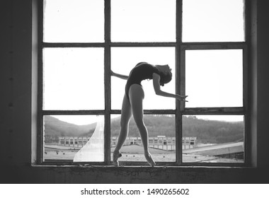 Black And White Photo Of Ballerina In The Window Frame In An Old Building. Young, Elegant, Graceful Woman Ballet Dancer