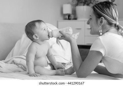 Black And White Photo Of Beautiful Smiling Mother Feeding Her 9 Months Old Baby From Bottle At Bedroom