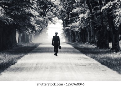 Black And White Photo Of A Businessman Or Gentleman In A Suit Walking Away On A Dusty Gravel Road. Business Man Walking In An Alley. Old Trees And Mysterious Leaving Of A Man In A Suit.