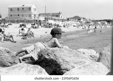 Black and white photo of a busy beach with buildings and a shirtless boy in a hat and sunglasses in the foreground laying on rocks and gazing into the ocean.