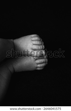 A black and white photo capturing the tiny feet of a newborn baby. The baby’s toes are curled and gently touching each other, set against a dark, soft background. The image highlights the softness 