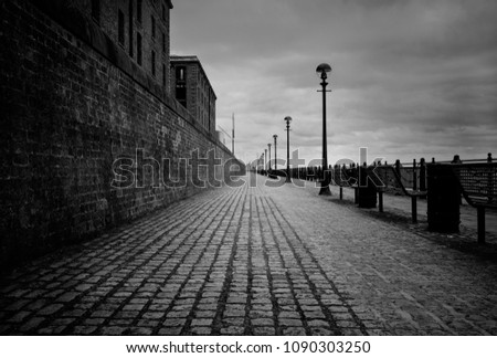 A black and white photo in the center of a cobblestone path on the Liverpool waterfront. The eerie sky fills the sky and a red brick wall aligns the left side of the photo.