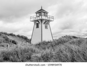 A Black And White Photo Of A Classic, 19th Century Wooden Lighthouse Towering Above Grassy Sand Dunes Under Large Storm Clouds.