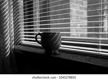 A Black And White Photo Of Coffee Mug Sitting On Window Ledge. The Window Blinds Are Open, Creating A Line Of Shadows On The Mug.