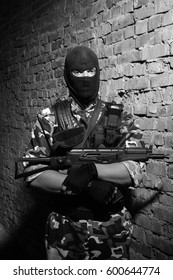 Black & White Photo Of Dangerous Terrorist Man In Black Balaclava Mask Holding Automatic Assault Rifle.Soldier In Heavy Combat Ammunition Posing In Ruined Building.Big Tough Guy With Gun In Black Mask