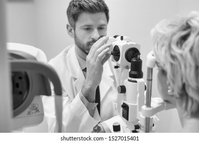 Black And White Photo Of Doctor Looking At Camera While Examining Senior Womans Eye At Hospital