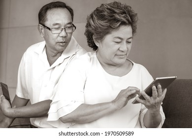 Black And White Photo Of Elderly Man Peeks Woman Cell Phone With Curiosity To Know. Senior Couple Stays Home Together. Older Male Takes A Secret Look At Wife Mobile Screen. Marriage Mistrust Concept