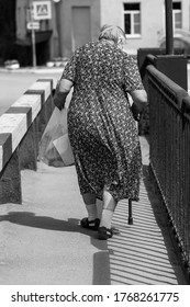 Black And White Photo Of An Elderly Woman Who Leaves A Store With Purchases On A Summer Day. An Elderly Overweight Woman Is Walking With A Heavy Bag And Wand.