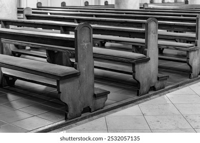 A black and white photo of empty wooden pews in a church. The pews are lined up in rows, and the floor is tiled. The pews are empty, and the light in the church is dim. - Powered by Shutterstock