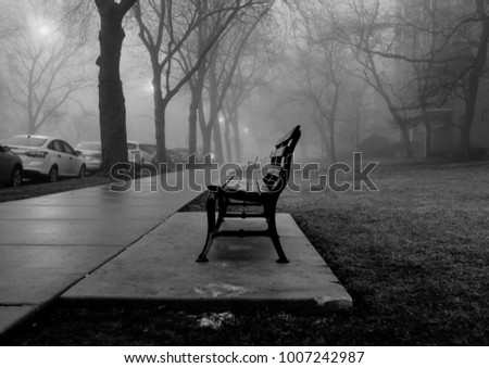 A black and white photo of a foggy city park bench during early winter. The trees are bare and the leaves that does the ground are dead.