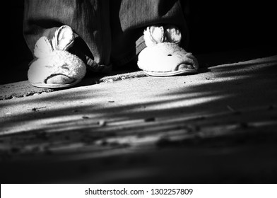 Black And White Photo Of Girl's Feet Wearing Sad Bunny Slipper House Shoes Sitting Alone Outside On Concrete Slab In A Low Income Housing Project One Easter Afternoon.