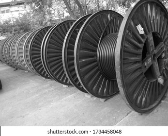Black and white photo of industrial cable reels in a diagonal line with trees behind. 