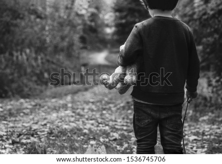 Black and white photo of Kid holding teddy waking alone in the forest, Rear view of a Boy standing alone holding his ted, Spoiled child, lost children or homeless kid concept