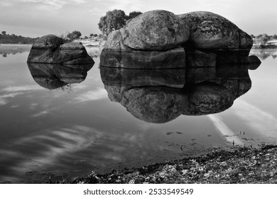 Black and white photo of large rocks reflecting in a calm lake with distant trees and a cloudy sky. - Powered by Shutterstock