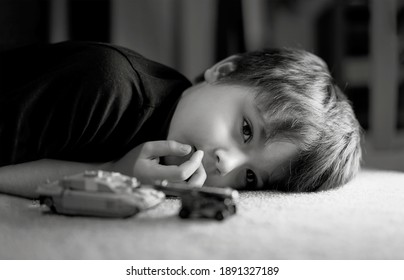 Black and white photo of lonely boy laying down on floor looking out deep in thought, Sad kid laying on carpet with tank toys.Child with bored face,Upset boy with unhappy face,Spoiled children