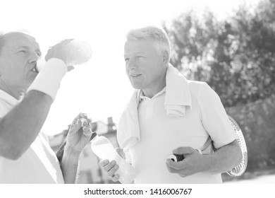 Black And White Photo Of Mature Man Looking Senior Friend Drinking From Bottle While Standing On Tennis Court During Summer