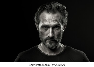 Black And White Photo Of Mid Aged Man Showing Severe Emotion. Beardy Male In Black T-shirt On Dark Background With Serious Look.