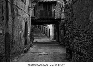 A black and white photo of a narrow, old street in a historic European town with stone buildings and a balcony. - Powered by Shutterstock