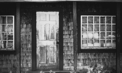 Black And White Photo Of An Old Dilapidated Shingled House Facade Including Front Door And Two Windows