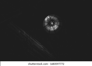 Black And White Photo Of Retro Metal Lampshade With Glass Light Bulb Inside. Glowing Vintage Streetlight Closeup On Black Background With Copy Space. Dramatic Lighting.