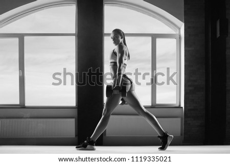 Black and white photo of sporty woman with strong body making exercises in spacy hall with panoramic windows. looks fit, stunning. powerful muscles. Keeping big dumbbells. Wearing stylish sportswear.